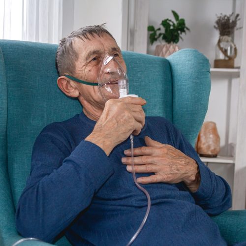 An elderly senior sits in a armchair with an oxygen mask in quarantine at home